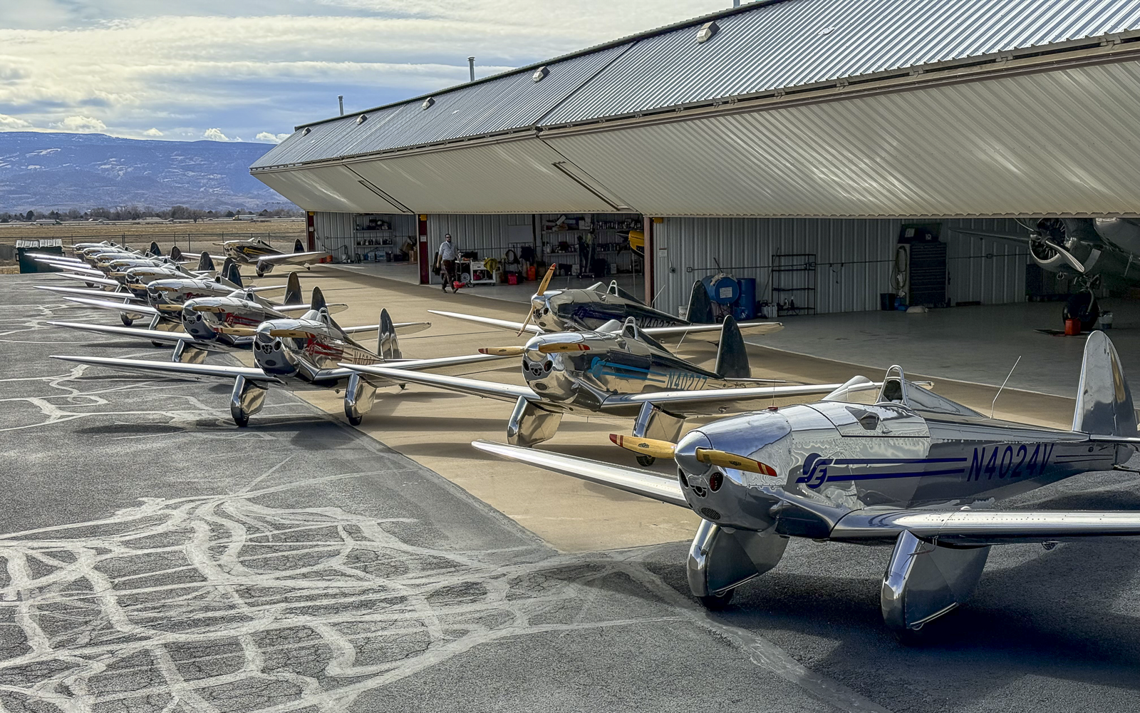 A row of polished silver vintage single-engine airplanes parked outside a long hangar, mountains in the distance.