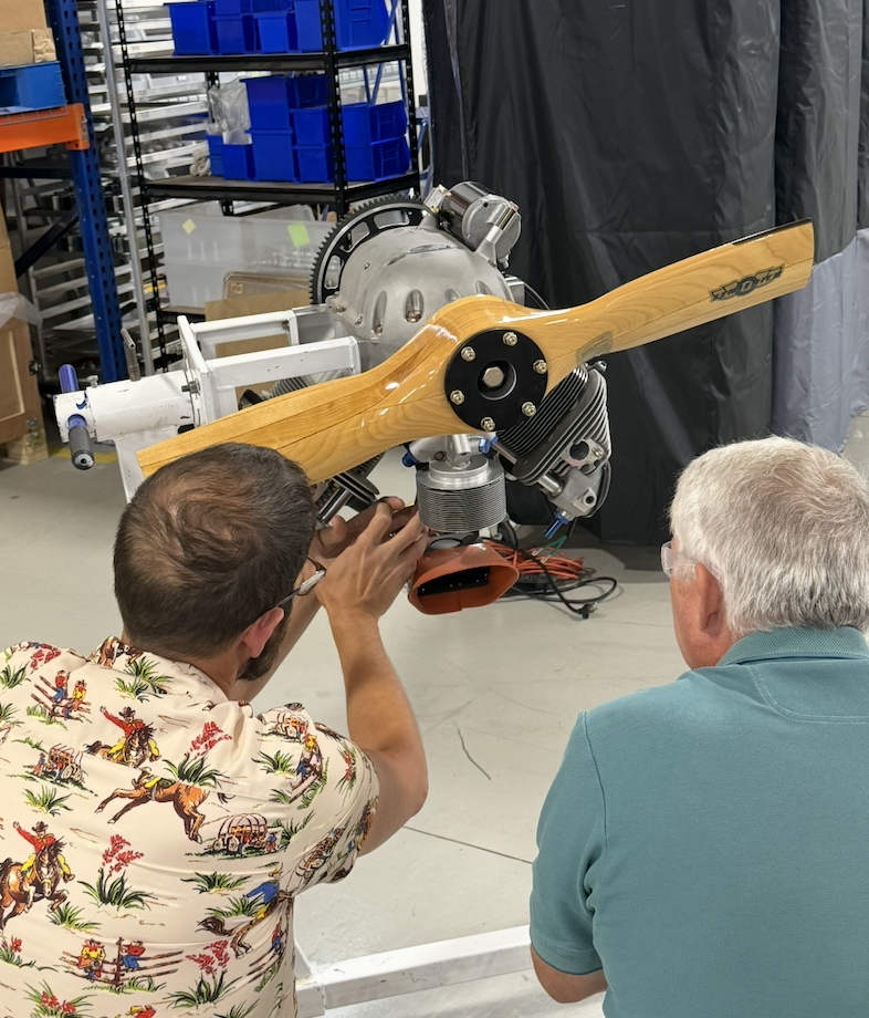 Two men examine a small aircraft engine with a wooden propeller, mounted on a stand in a workshop.